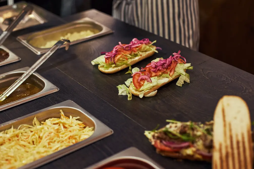 Sandwiches being assembled with lettuce and pickled red onion, next to trays of prep ingredients